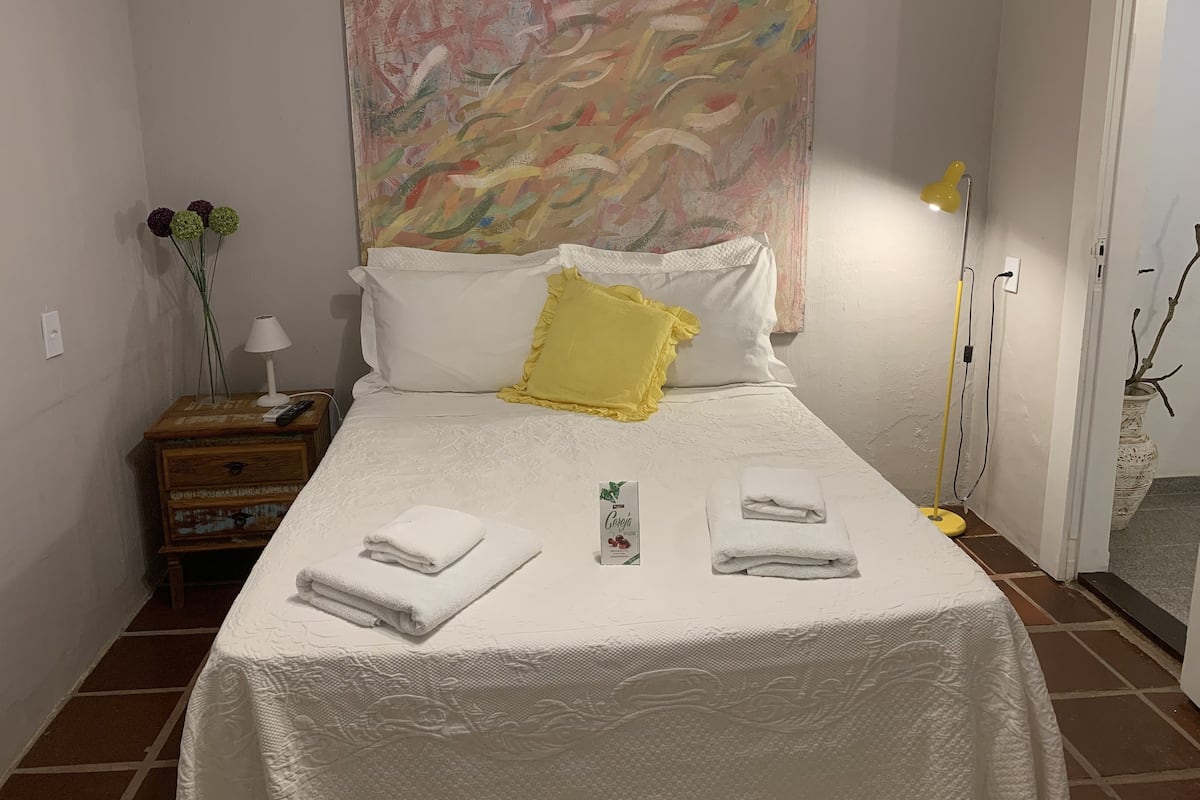 Un lit bien fait, avec du linge de lit blanc et un coussin jaune, une table de chevet avec une lampe et des fleurs, et un grand tableau abstrait accroché au mur.