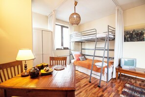 Une petite pièce comprenant un lit superposé, une table en bois avec une lampe, une petite télévision et un tableau représentant un paysage urbain accroché au mur.