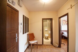 Un couloir avec une porte en bois, une chaise sur laquelle est posée une serviette, une salle de bain avec des toilettes et un lit dans la pièce adjacente.