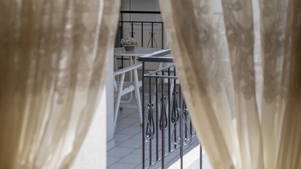 Blick durch leichte Vorhänge auf einen Balkon mit Tisch, Stühlen und Geländer.