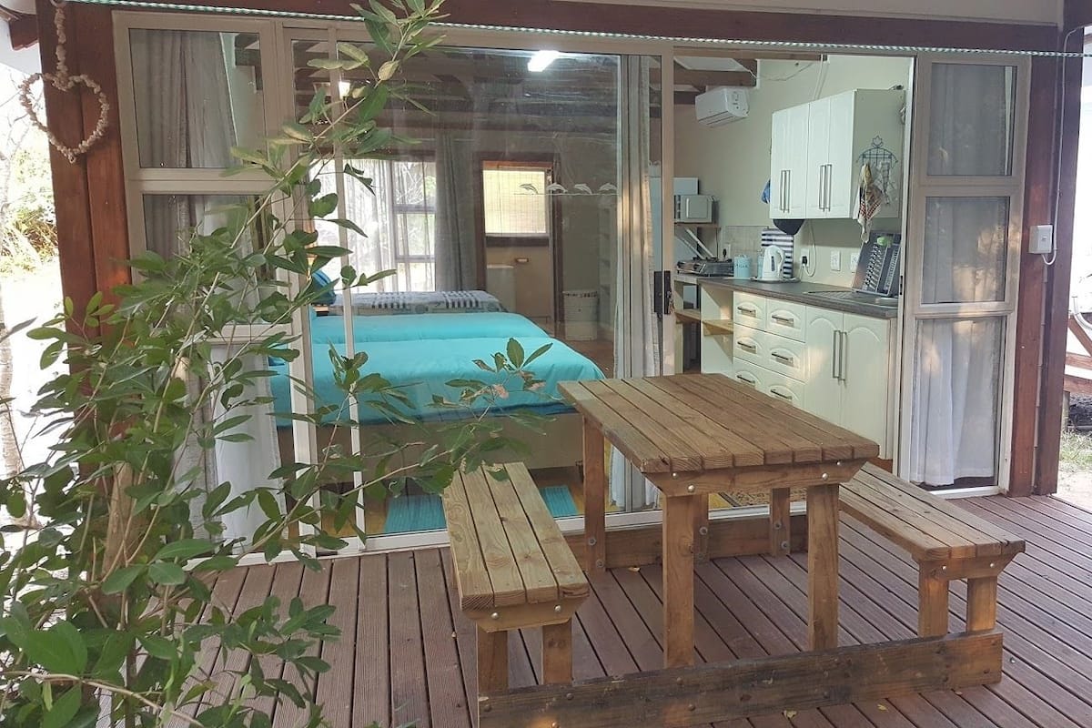 Een houten terras met een tafel en banken, een schuifdeur naar een slaapkamer met een bed en een kitchenette.