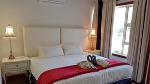 Una cama bien hecha con una manta roja y dos toallas dispuestas en forma de cisne, almohadas blancas y un cabecero blanco. Hay dos lámparas de noche en mesitas blancas, y una ventana con cortinas a la derecha.