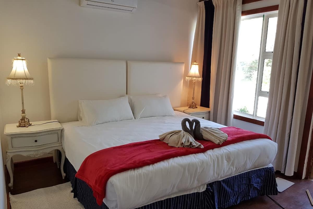Een net opgemaakt bed met een rode deken en twee handdoeken gevouwen in de vorm van een zwaan, witte kussens en een witte hoofdbord. Twee bedlampen staan op witte nachtkastjes, en aan de rechterkant is een raam met gordijnen te zien.