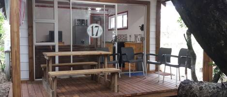 Una terraza de madera con una mesa y sillas de comedor, un área de cocina con ventana, y una puerta con un cartel de número.