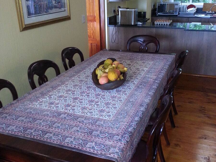Comedor con una larga mesa, sillas y un cuenco de fruta. Se observa una cocina al fondo con electrodomésticos y una ventana.