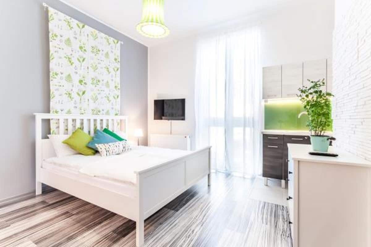 A modern bedroom with a white bed, a small desk, and a kitchen area with white cabinets.