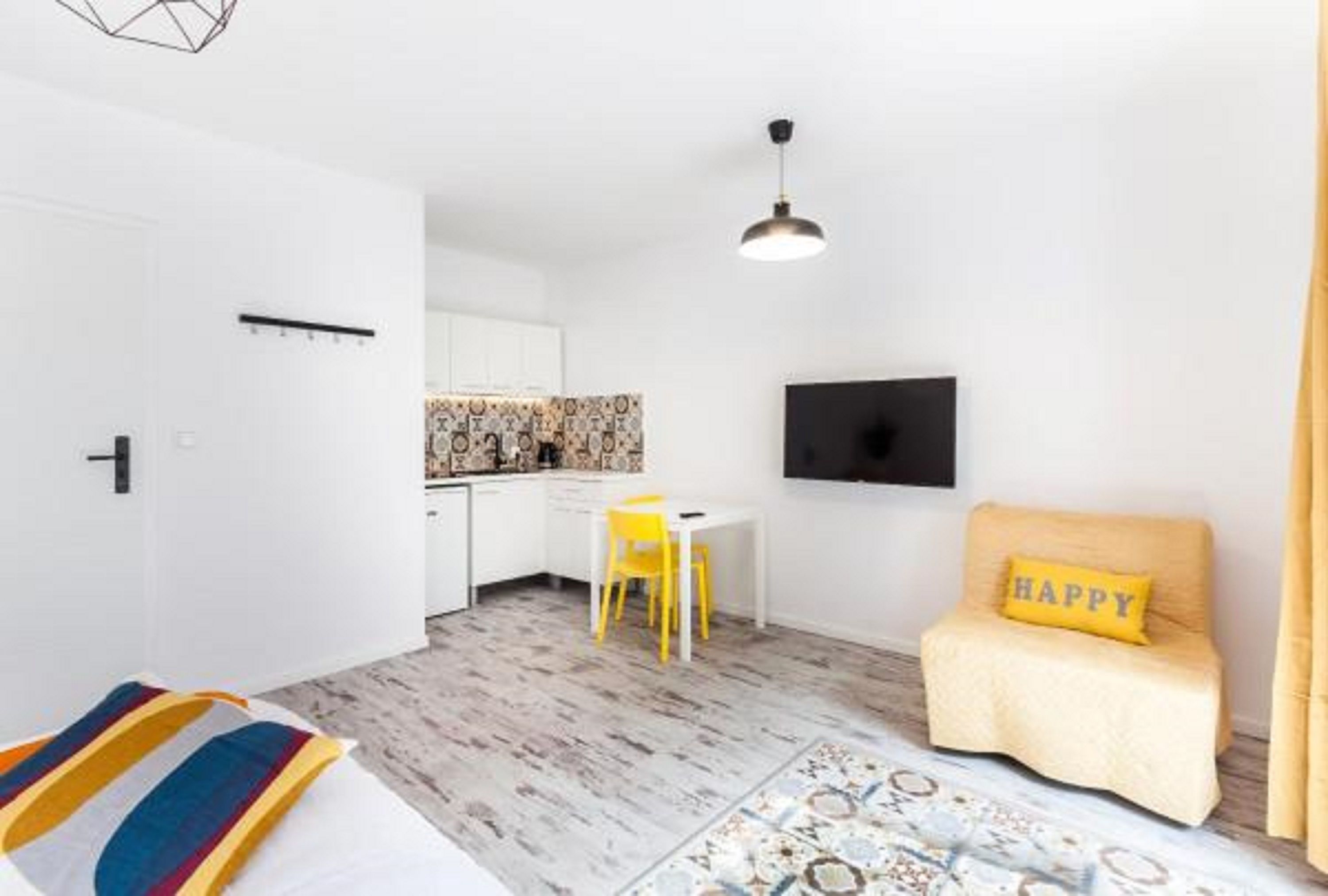 A compact hotel room with a bed, a small desk, a chair, a TV, and a sofa with a 'HAPPY' pillow.
