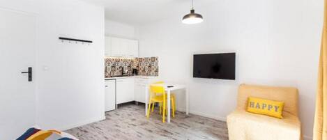 A compact hotel room with a bed, a small desk, a chair, a TV, and a sofa with a 'HAPPY' pillow.