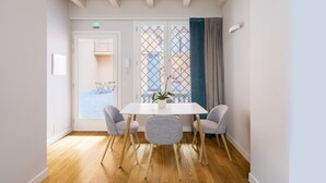 A dining area with a white table, four grey chairs, a potted plant, and a view through a window with a curtain. - Residenza Palline by Hili (Rome)
