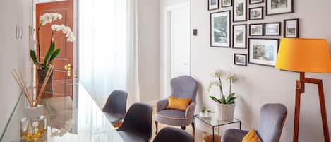 A modern living room with a glass coffee table, four grey armchairs, a small side table with a plant, and a lamp.