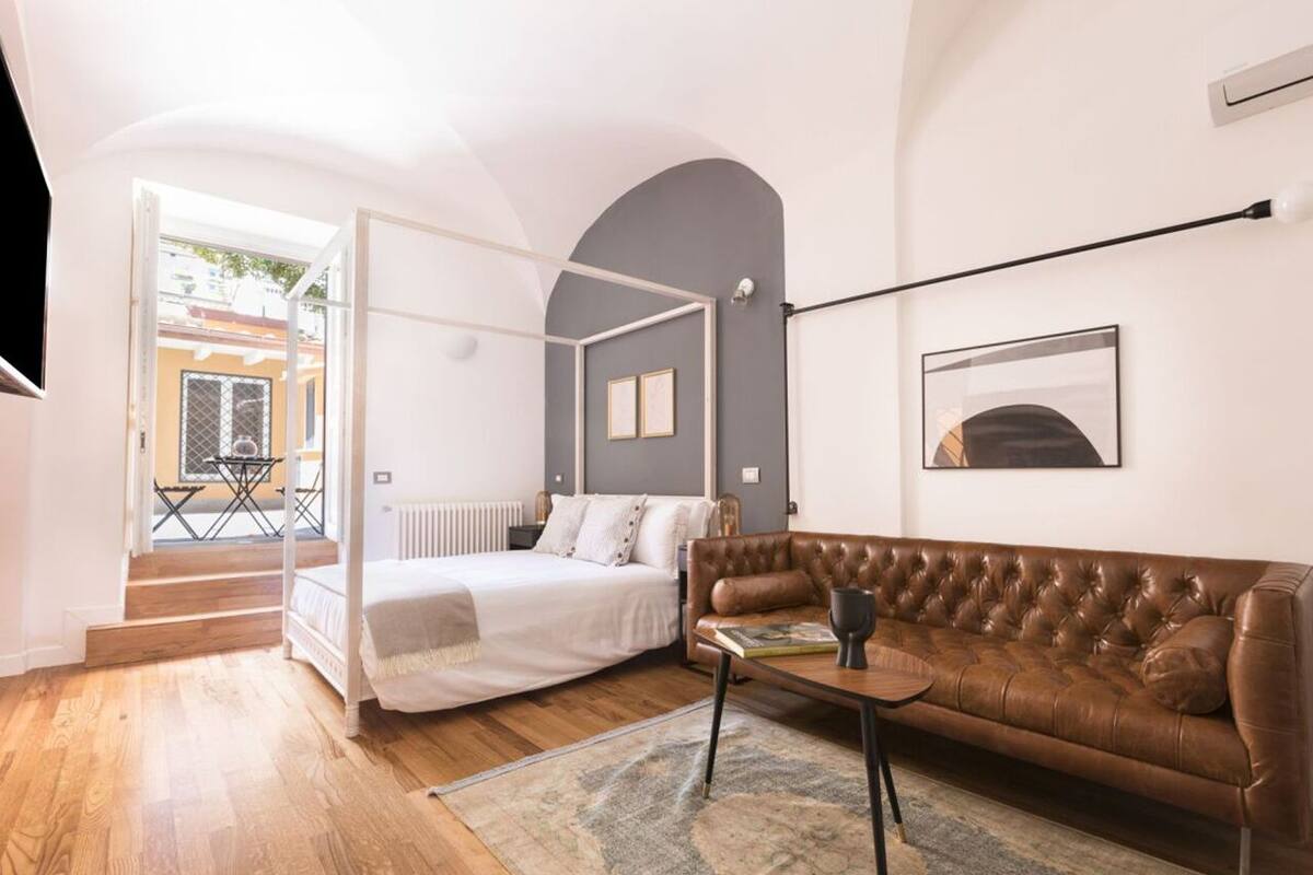 A bedroom with a bed, a brown leather sofa, a small table, and a view of the outside.