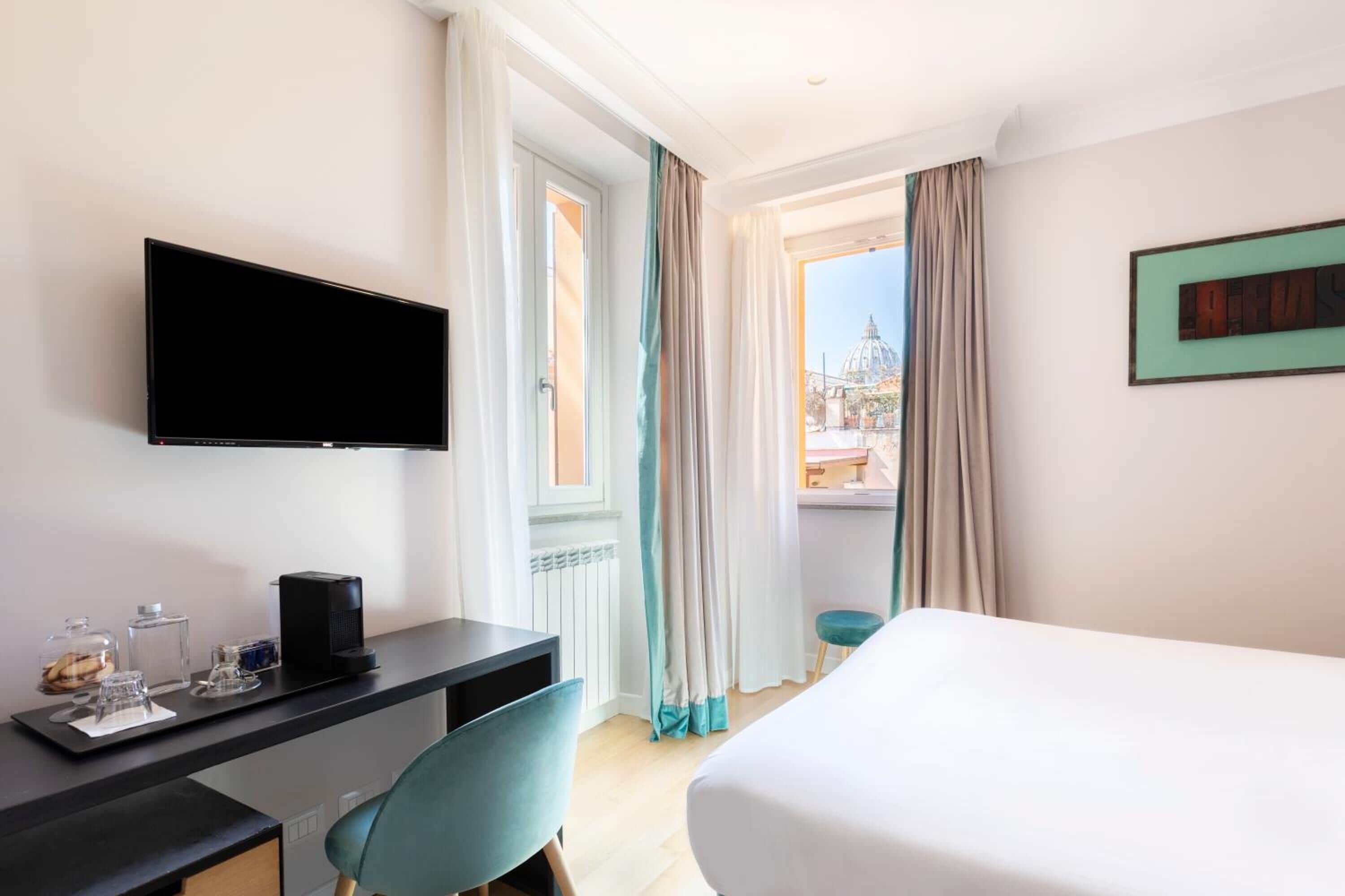 A hotel room with a bed, a desk with a computer, a TV, and a window with curtains.