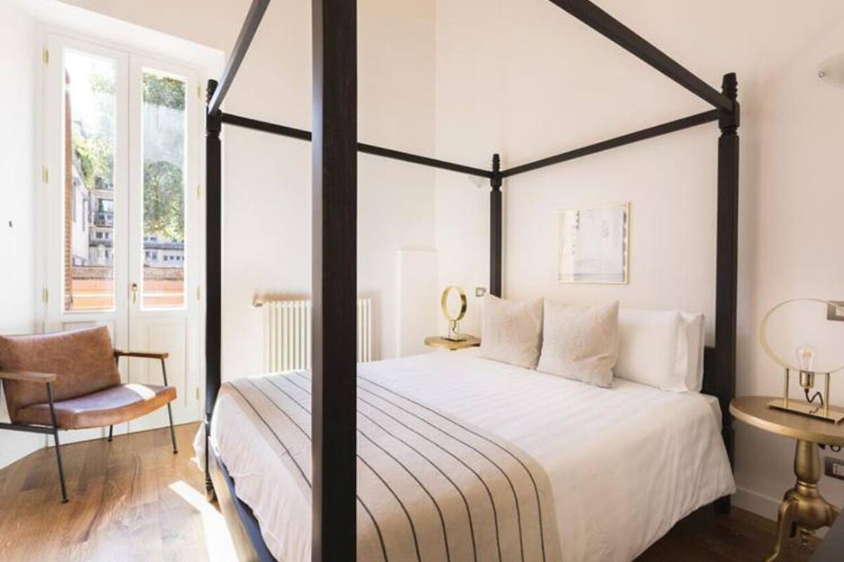 A four-poster bed with a striped bedspread, a chair, a bedside table with a lamp, and a view of the outside through a door.