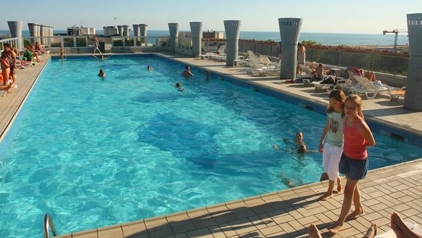 A swimming pool with people swimming and lounging, surrounded by sunbeds and a view of the sea. - Condominio Terme (San Michele al Tagliamento)