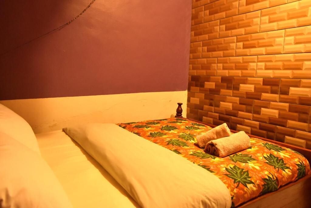 A single bed with a patterned bedspread and two pillows, a wooden headboard, and a plain wall.