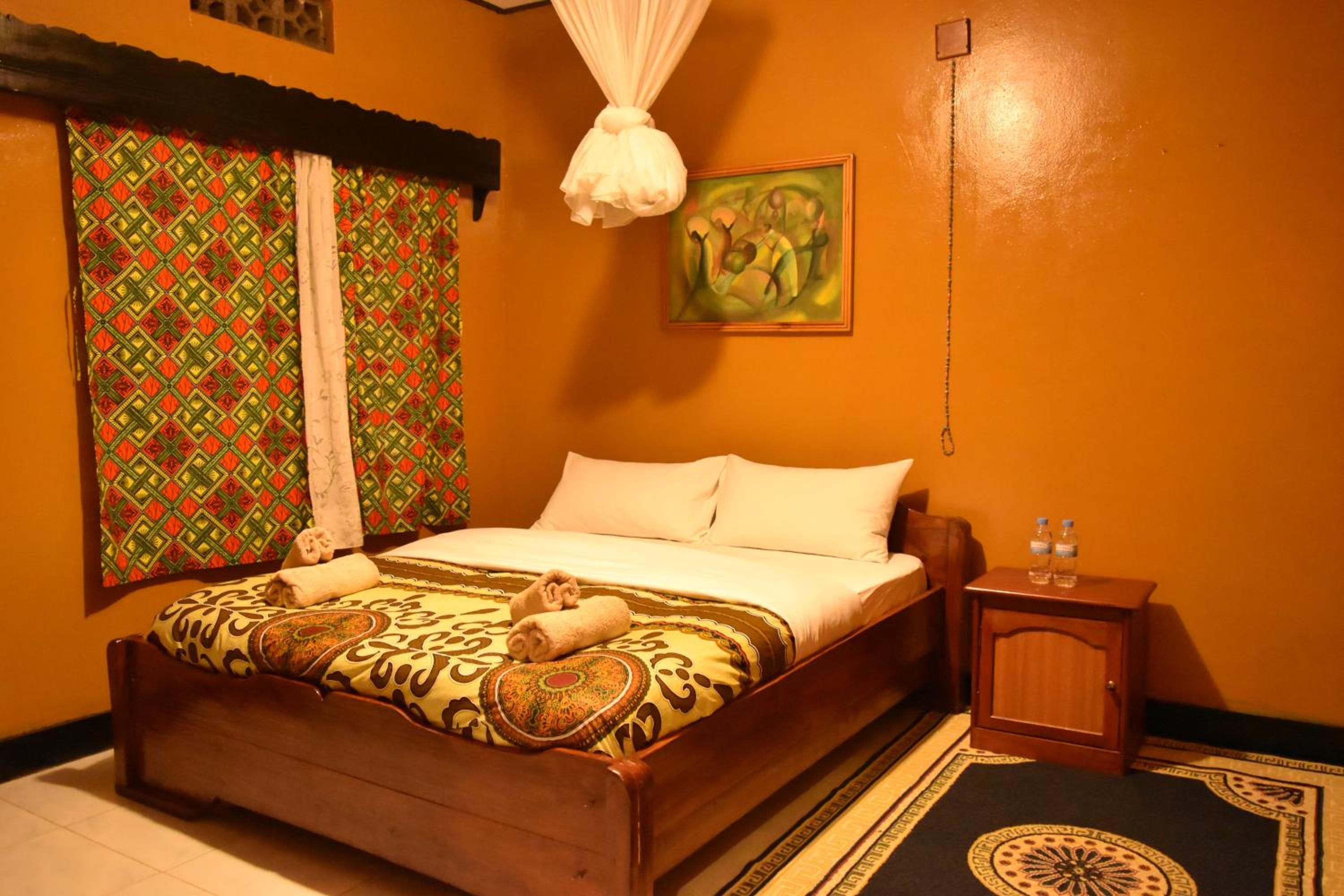 A hotel room with a wooden bed, patterned bedspread, a small wooden nightstand with water bottles, and a painting on the wall.