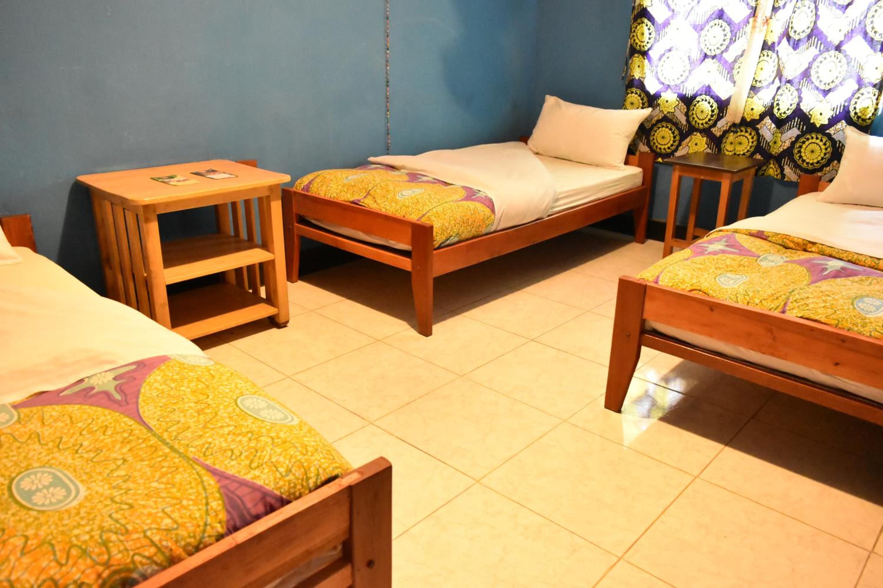 A room with three wooden beds, each with a patterned bedspread. There is a small wooden table with a chair, and a window with patterned curtains.