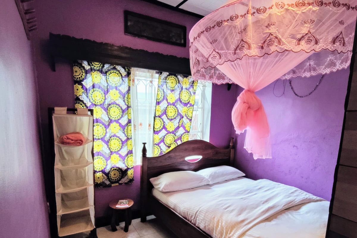 A bedroom with a bed, a window with patterned curtains, a bedside table, and a shelf unit.