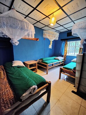 A room with two wooden beds, each with green bedding and white pillows. There are two bedside tables with lamps. The room has a blue wall and a window with patterned curtains. - Amahoro Guest House (Muhoza)