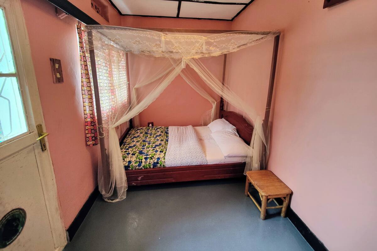 A room with a bed under a canopy, a small wooden stool, and a window with patterned curtains.