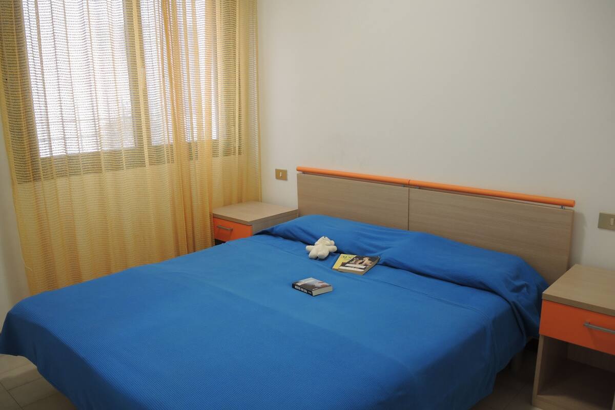Een netjes opgemaakt bed met een blauw sprei, een klein nachtkastje en een bijzettafeltje met een lamp.