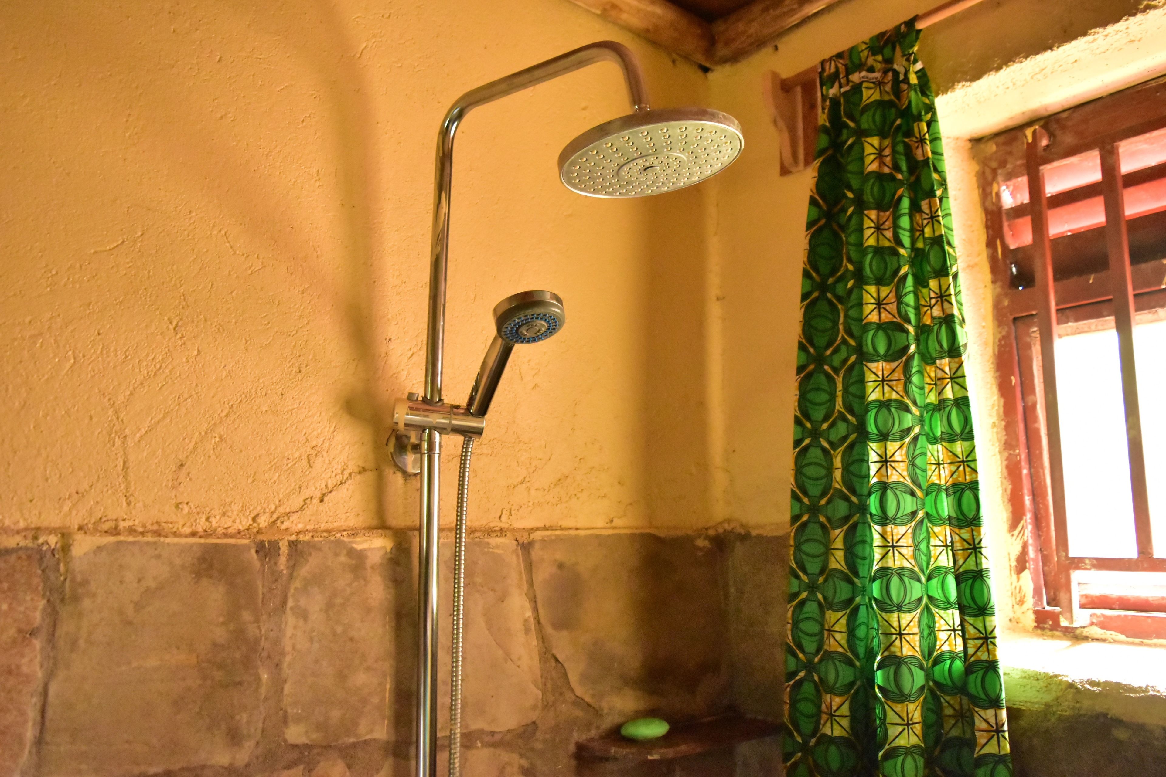 Een douche met een handdouche en een vaste douchekop, een gordijn met een groen patroon, en een stenen muur.