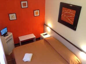 A hotel room with a bed, a desk with a computer, a bedside table with a lamp, and framed artwork on the wall. - Hotel Arco del Sole (Lavagna)