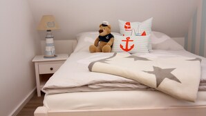 Un llit ben fet amb una manta de motius estrellats, un os de peluix amb gorro de mariner, almoines amb ratlles i una làmpada de far a la taula de nit.