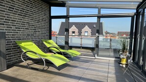 Un balcón soleado con dos sillones verdes, un ventanal de vidrio que da a una terraza y una planta en maceta.