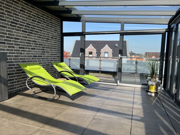 A sunlit balcony with two green lounge chairs, a glass door leading to a terrace, and a potted plant.