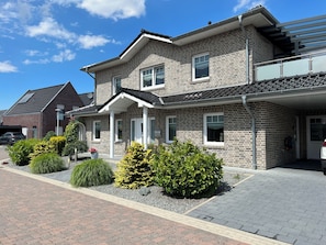 A two-story residential building with a brick facade, a covered porch, and a driveway with landscaped bushes.