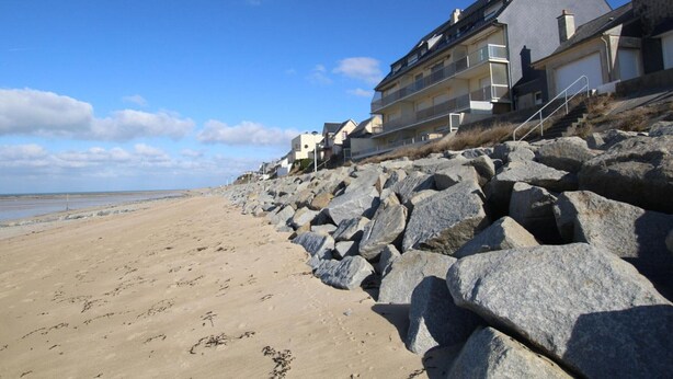 Ein Strand mit großen Felsen im Vordergrund und einer Reihe von Gebäuden mit Balkonen im Hintergrund.