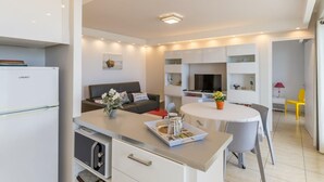 Eine moderne Küche mit integriertem Kühlschrank, ein Essbereich mit rundem Tisch und Stühlen sowie ein Wohnbereich mit Sofa und Fernseher.