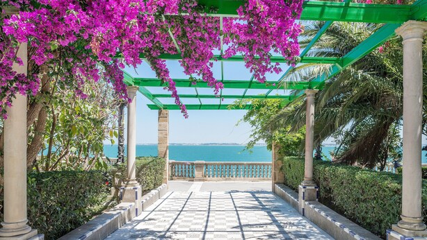 Un sentier menant à un point de vue sur la mer, agrémenté d’une pergola recouverte de fleurs roses et de colonnes.