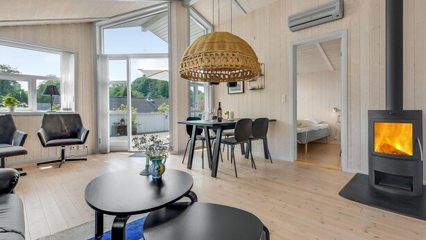 En moderne stue med et stort spisebord, behagelige siddepladser, en pejs og store vinduer med udsigt til naturen.