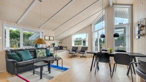 En rummelig opholdsstue med sofa, sofabord og spisebord med stole. Store vinduer giver naturligt lys og udsigt til grønne omgivelser.