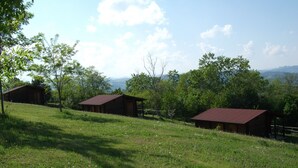 Un paysage rural avec trois cabanes en bois sur une colline verdoyante, entourées d’arbres et avec des montagnes en arrière-plan.