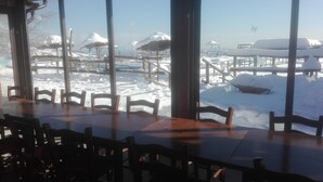 Un espace repas offrant une vue sur un paysage enneigé, avec notamment une terrasse en bois et des tables et des chaises recouvertes de neige.