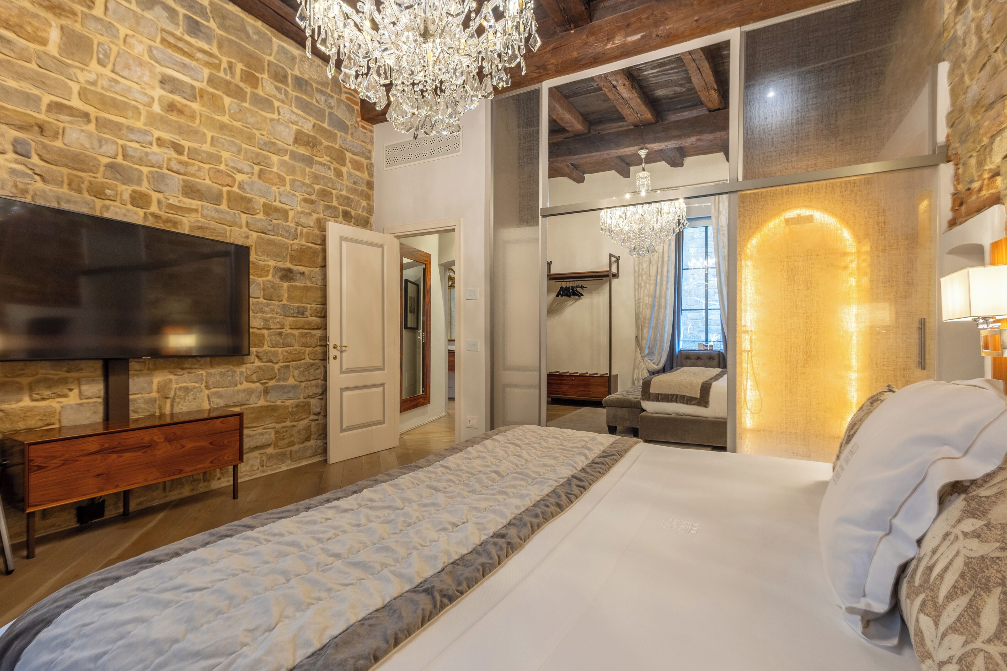 Une chambre avec un mur en pierre, un téléviseur à écran plat, un banc en bois, un grand lit, un miroir et un lustre.