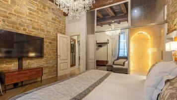 Une chambre avec un mur en pierre, un téléviseur à écran plat, un banc en bois, un grand lit, un miroir et un lustre.