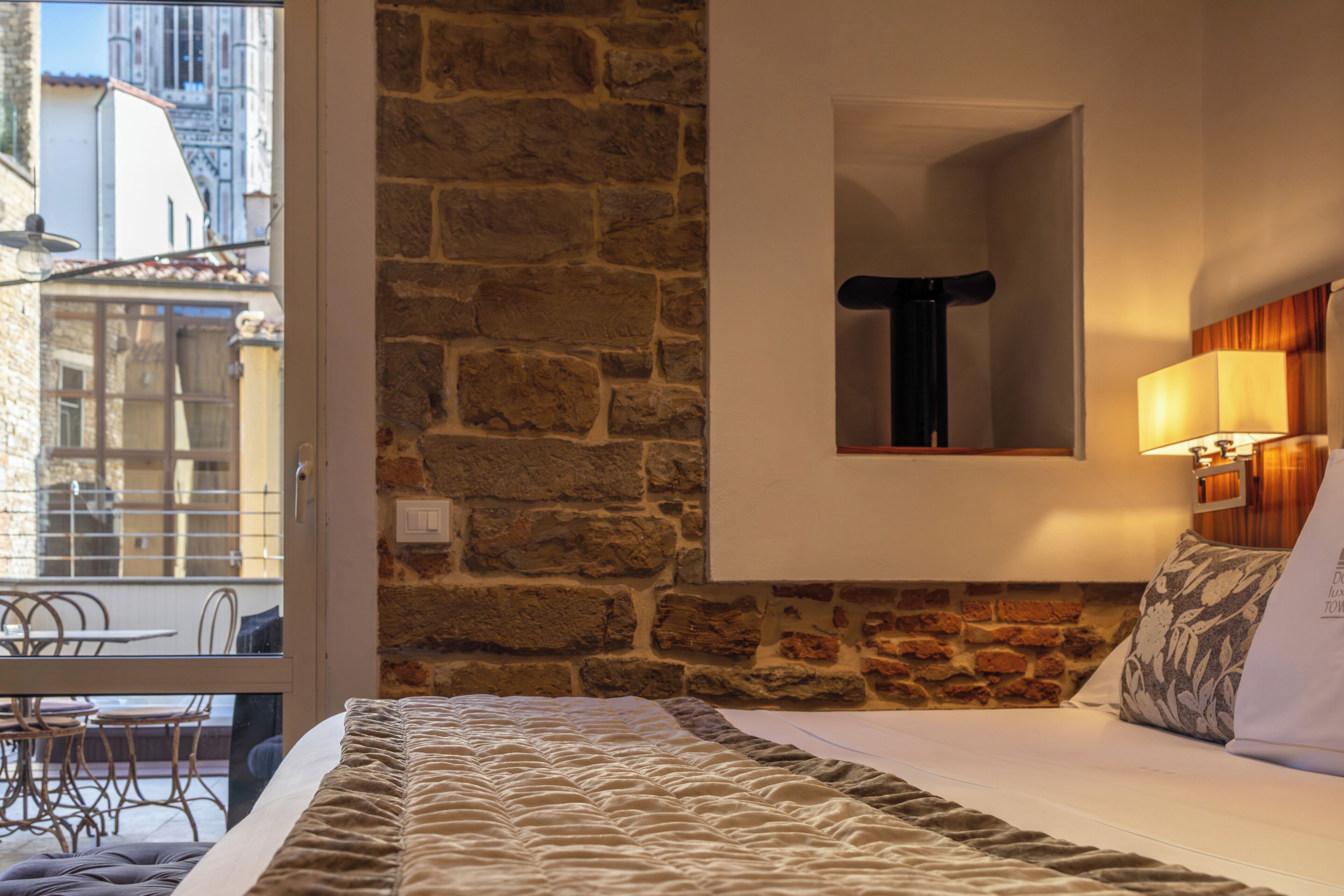 Quarto de hotel com parede de pedra, uma cama com edredom branco, uma mesinha e uma cadeira.