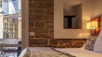Quarto de hotel com parede de pedra, uma cama com edredom branco, uma mesinha e uma cadeira.