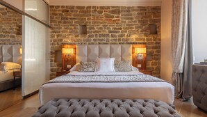 Quarto com parede de pedra, uma cama grande com cabeceira estofada, duas mesas de cabeceira com luminárias e um banco estofado cinza.