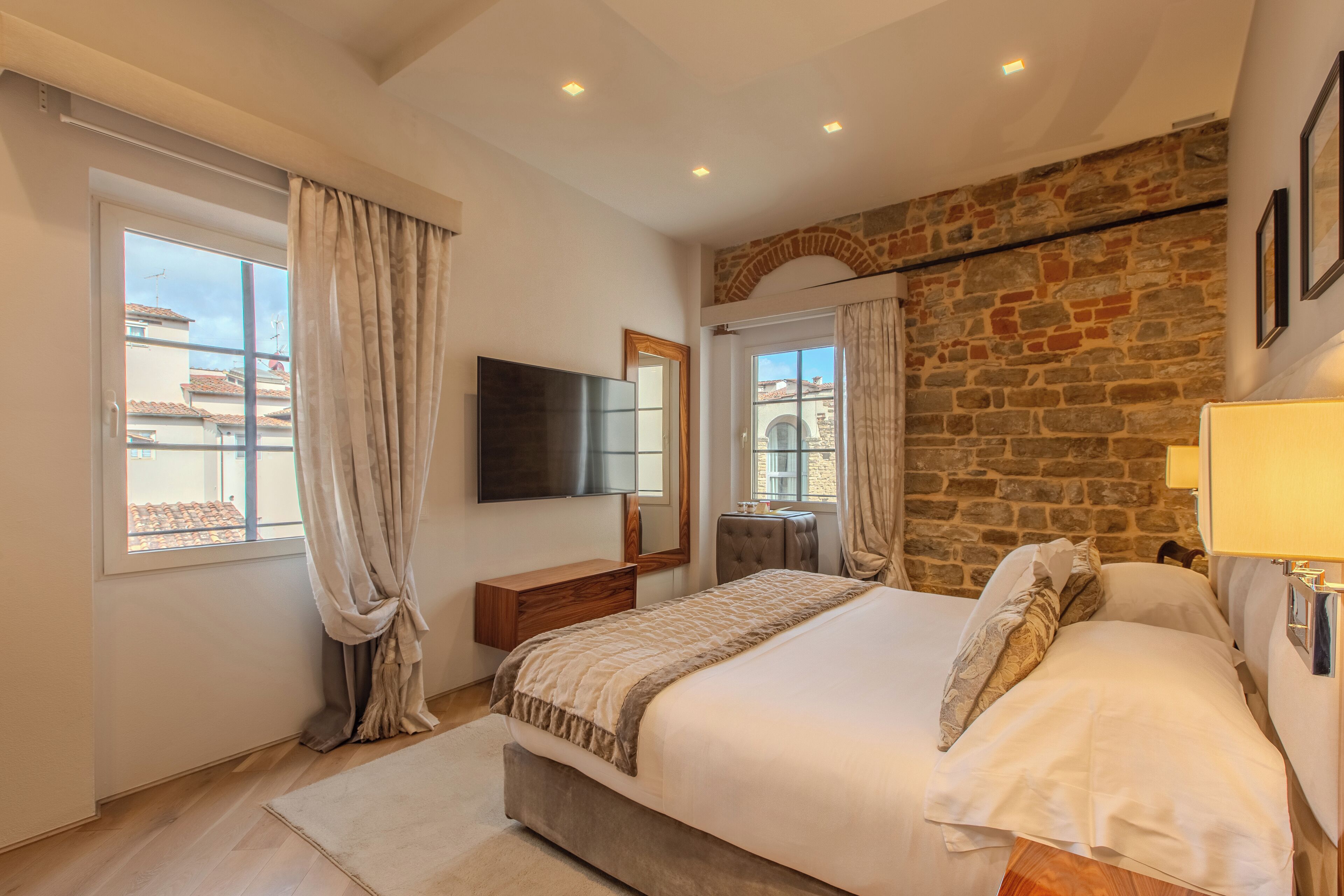 A bedroom with a large bed, a flat-screen TV, a stone wall, and a window with curtains.