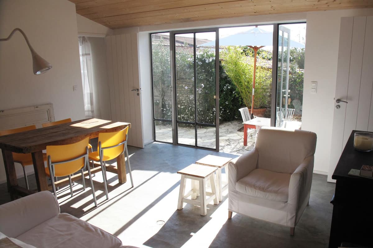 Ein großzügiger Wohnbereich mit Esstisch, gelben Stühlen, einem weißen Sofa und einem beigefarbenen Sessel. Eine Glastür führt auf eine Terrasse mit Blick auf Grünflächen und ein Gebirge.