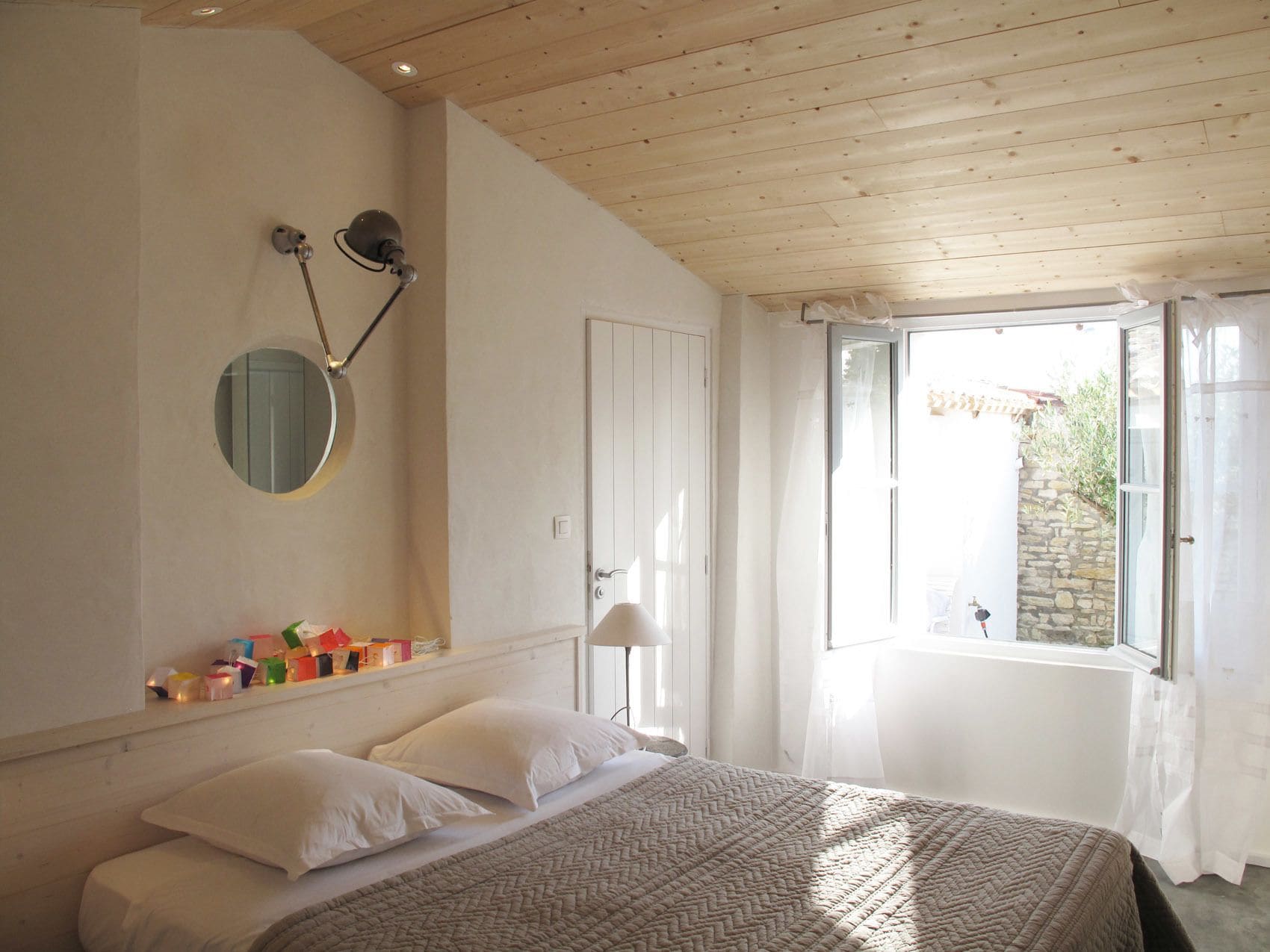 Camera da letto con un letto, uno specchio tondo, un comodino con diversi oggetti, una finestra con tende e vista all'esterno.