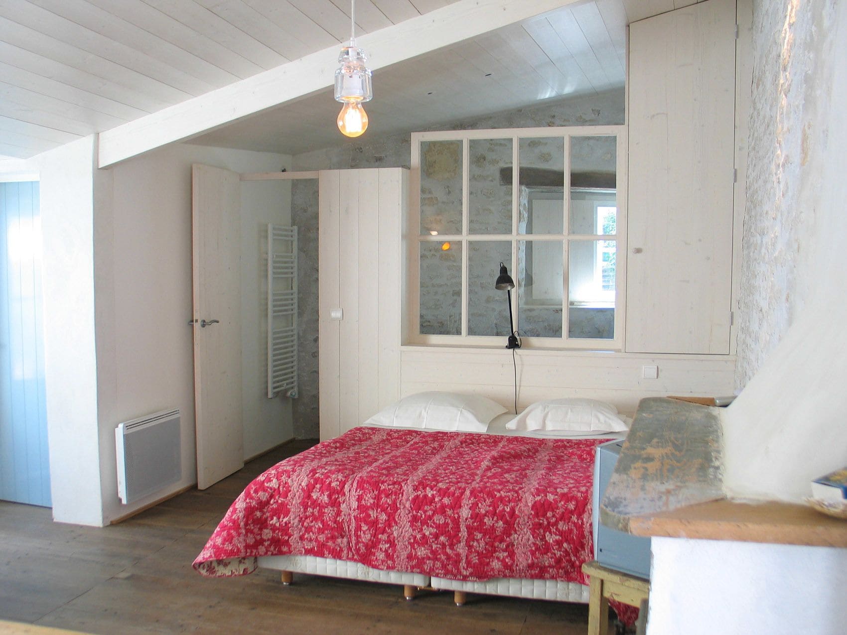 Une chambre avec un plafond en bois, une grande fenêtre, un lit recouvert d’une courtepointe à motifs floraux rouges et un radiateur.