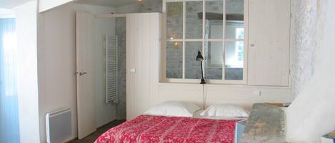 Une chambre avec un plafond en bois, une grande fenêtre, un lit recouvert d’une courtepointe à motifs floraux rouges et un radiateur.