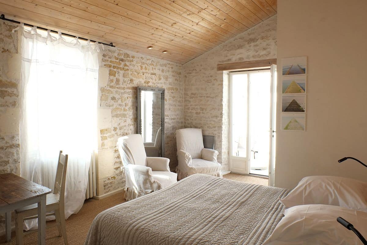 Ein Schlafzimmer mit Holzboden, Steinwänden, einem Bett mit einer beigefarbenen Decke, zwei weißen Stühlen und einem großen Spiegel.