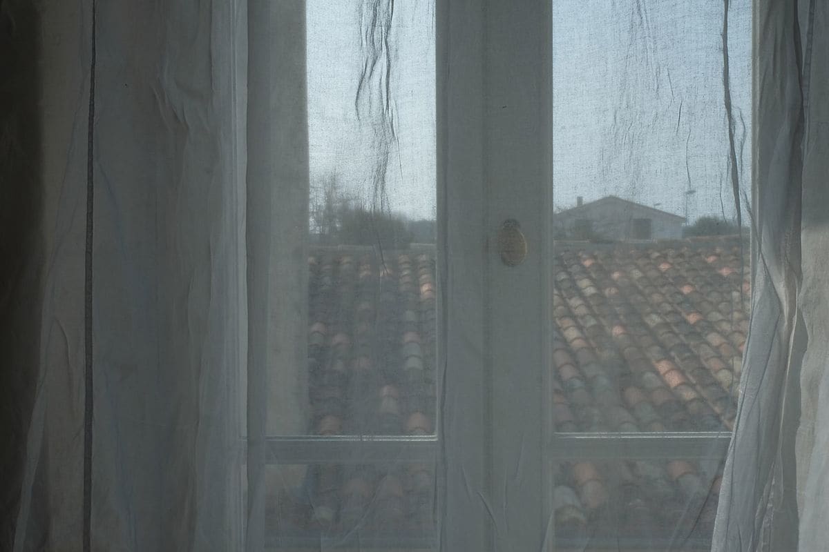 A view through a window with a metal grid, a door with a brass handle, and a tiled roof visible outside.